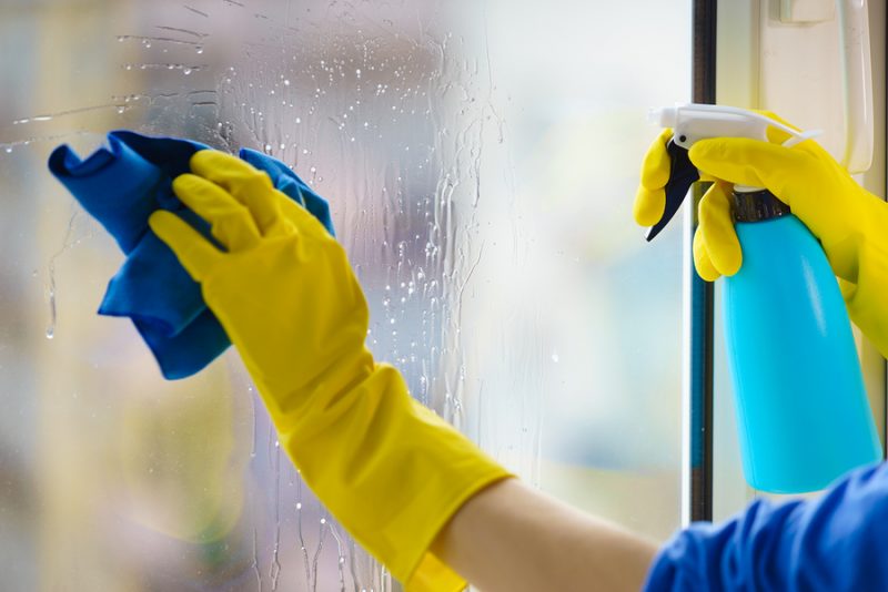 Female hand in yellow gloves cleaning window pane with rag and spray detergent. Cleaning concept